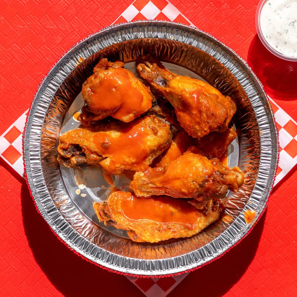 Buffalo wings in a foil container with ranch dressing on a red table with a checkered napkin.