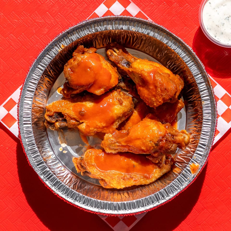 Buffalo wings in a foil container with ranch dressing on a red table with a checkered napkin.