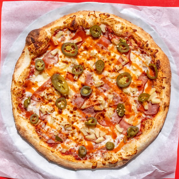 Whole pizza with ham, pineapple, jalapeños, cheese, and hot sauce drizzle on a red background.