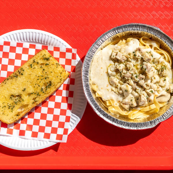 Garlic bread on a checkered paper and chicken Alfredo pasta in a foil container on a red tray.