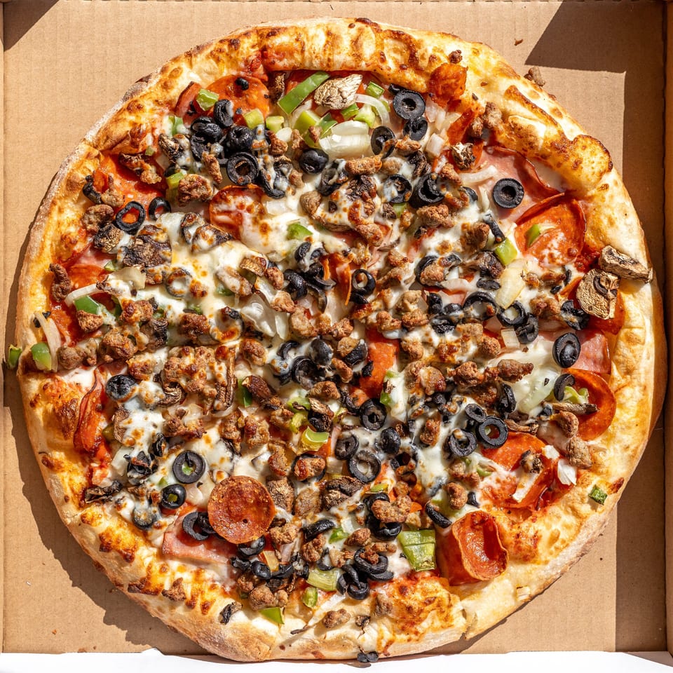 Overhead view of a loaded pizza with pepperoni, sausage, olives, peppers, and mushrooms in a box.