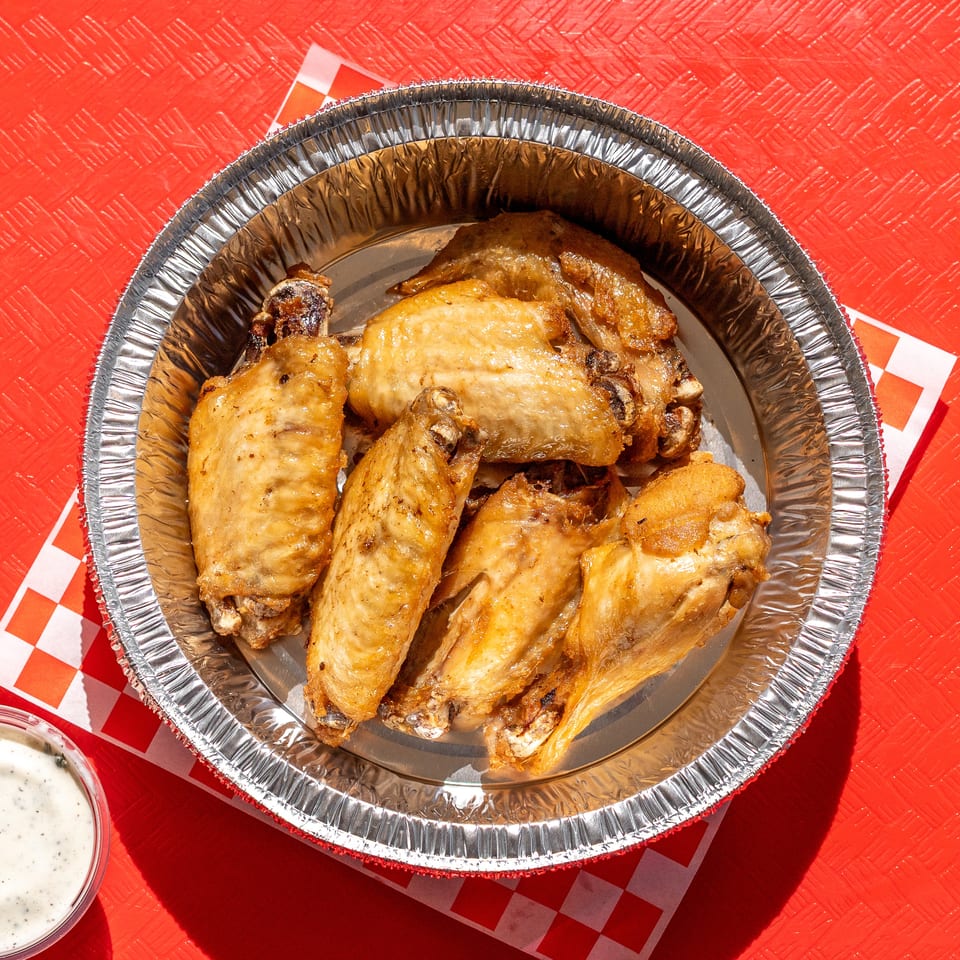 Golden brown chicken wings in a foil container with a side of ranch on a red checkered paper.