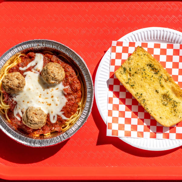 Spaghetti with meatballs and melted cheese in a foil container, with garlic bread on a red tray.