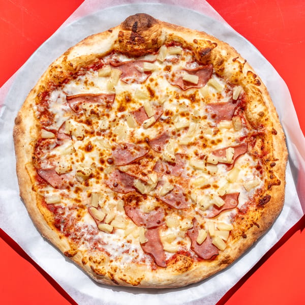 Hawaiian pizza with ham and pineapple on a white paper-lined tray against a red background.
