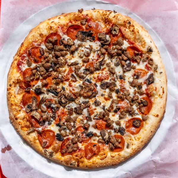 Whole pizza with pepperoni, sausage, and mushrooms on a metal tray over a red surface.