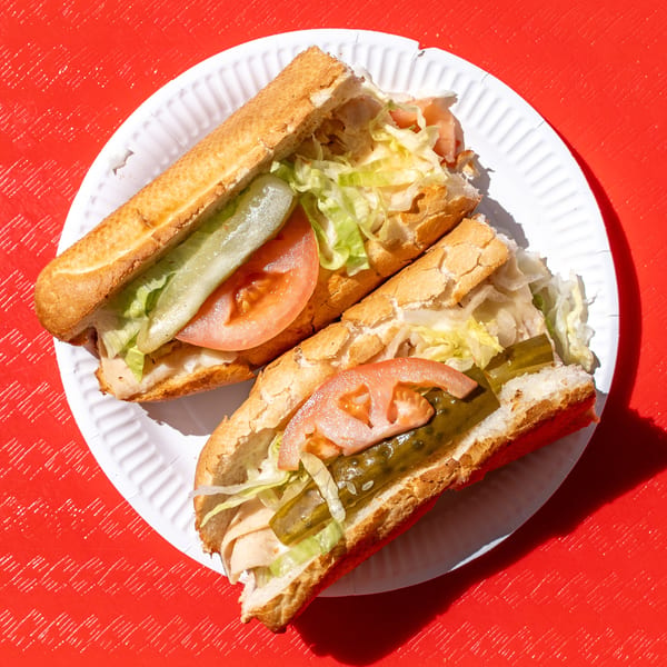 Two subs filled with lettuce, tomato, pickles, and turkey on a white paper plate.